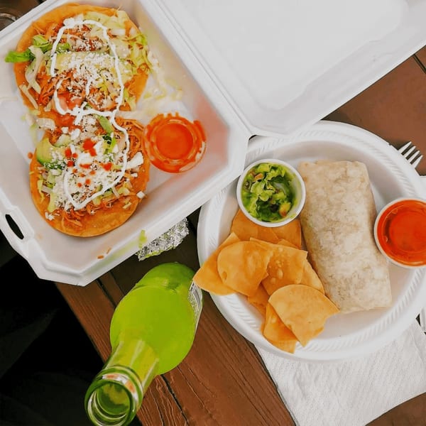Two El Oasis tostadas, a burrito with chips and guacamole, and a green soda bottle on a wooden table.