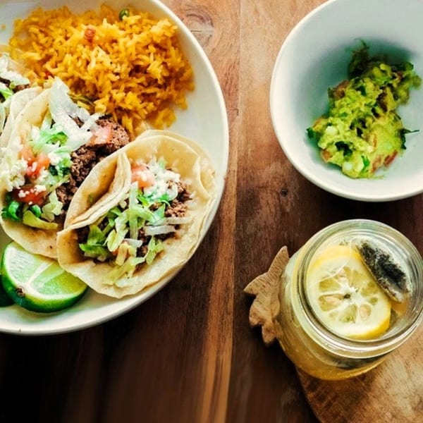 Three El Oasis tacos with rice, lime, guacamole, and a lemon drink on a wooden table.
