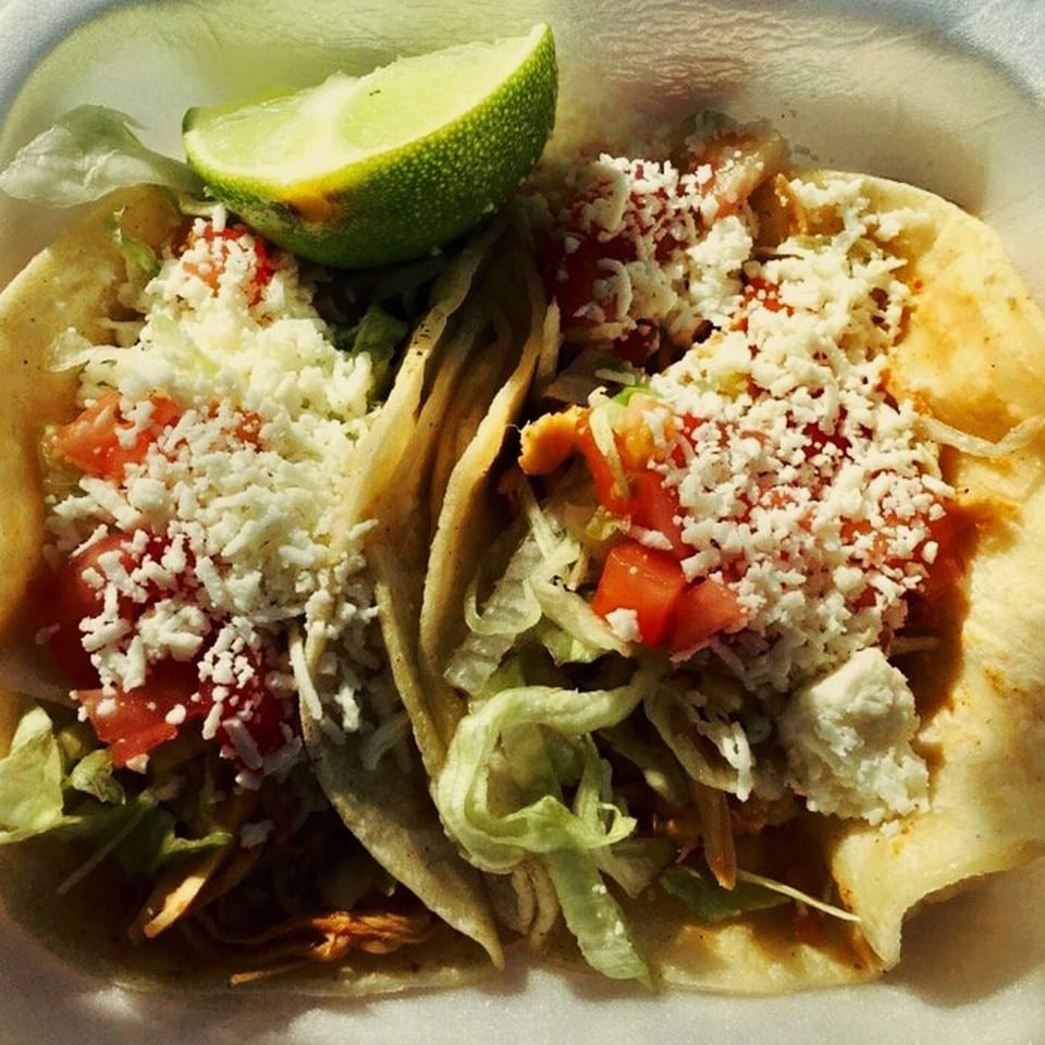 Two El Oasis tacos filled with meat, lettuce, diced tomatoes, and crumbled white cheese, with a lime wedge.
