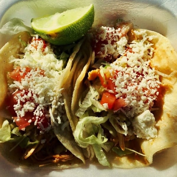 Two El Oasis tacos filled with meat, lettuce, tomatoes, and cheese, garnished with a lime wedge.